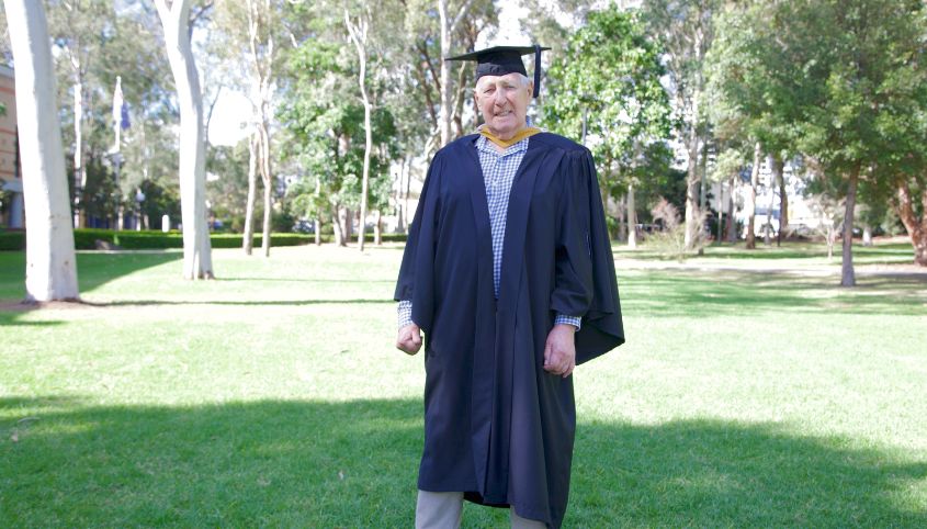 Roger Whalan stands in his graduation gown in front of  gum trees at Macquarie University